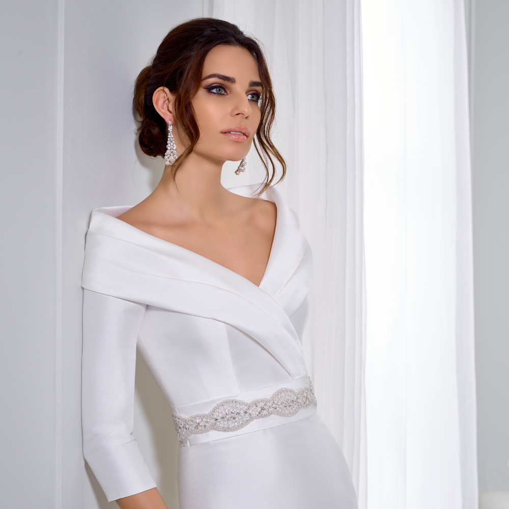 Close up of a brunette model stood against a wall in an off the shoulder white Mikado wedding dress with a lapel neckline and sparkle belt 